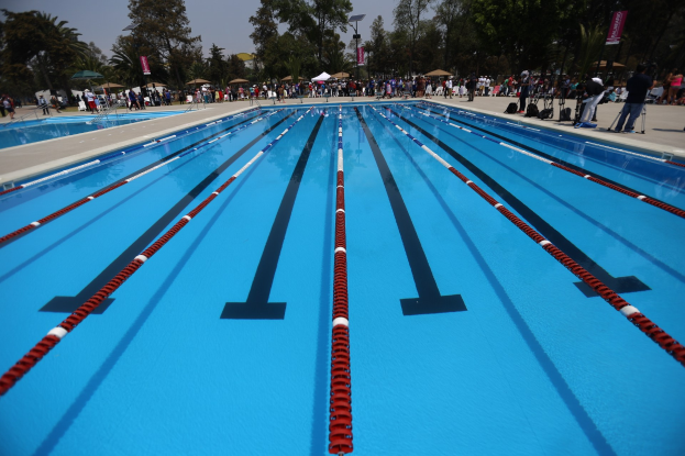 Großer Schwimmbad mit Bahnentrennungen, umgeben von Menschen mit Taschen und Gegenständen, mit Zelten, Schildern, Stangen, einer Tafel und einem bewölkten Himmel im Hintergrund.