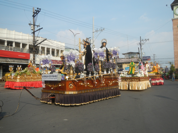Ein Paradewagen mit Menschen darauf, umgeben von Gebäuden, Strommasten, Laternenmasten, Bäumen und einem Uhrenturm im Hintergrund, unter einem bewölkten Himmel.