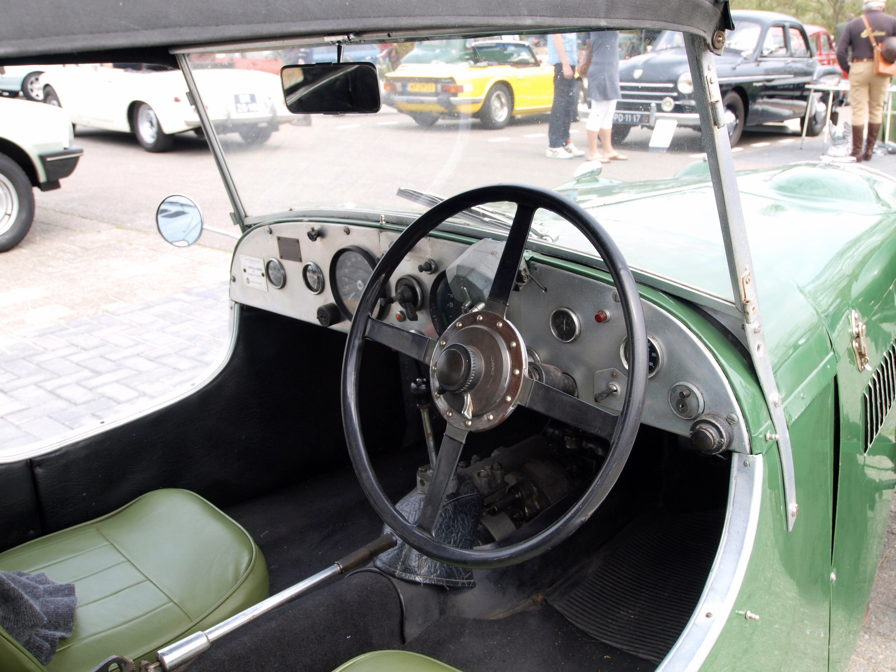 Innenansicht eines grünen Autos mit Lenkrad, Armaturenbrett, Sitzen und Rückspiegel, mit anderen Fahrzeugen, Fußgängern, Bäumen und einem blauen Himmel im Hintergrund.