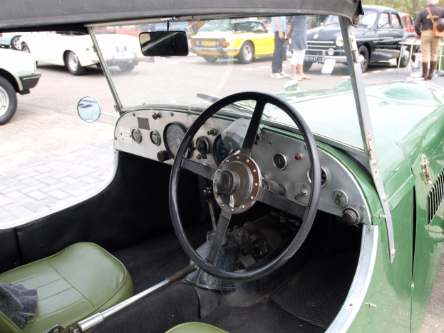 Innenansicht eines grünen Autos mit Lenkrad, Armaturenbrett, Sitzen und Rückspiegel, mit anderen Fahrzeugen, Fußgängern, Bäumen und einem blauen Himmel im Hintergrund.