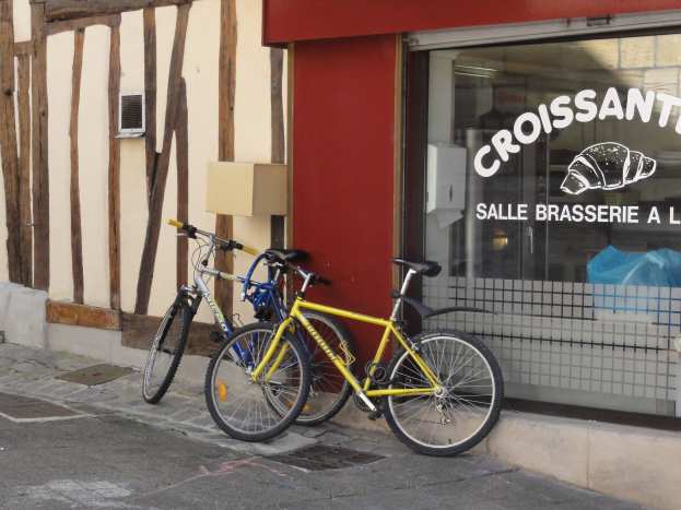 Zwei Fahrräder nebeneinander auf einem Gehweg vor einem Geschäft mit einer Glaswand, die Text enthält und ein Objekt links von den Fahrrädern.