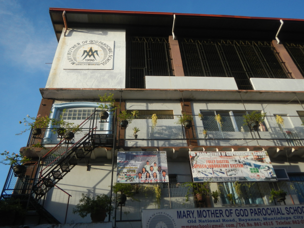 Das Marien-Mutter-Gottes-Grundschulgebäude mit Fenstern, Geländern, Treppen, Topfpflanzen, Bannern und einem bewölkten Himmel im Hintergrund.