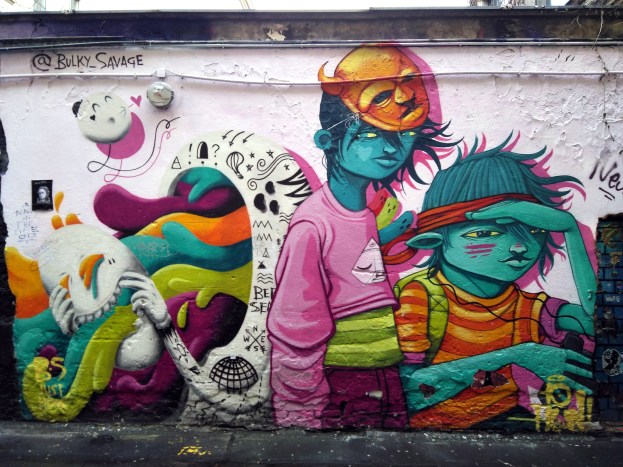 Mural an einer Gebäudeseite mit Graffiti, darunter eine Straße, Strommasten und einen klaren blauen Himmel.