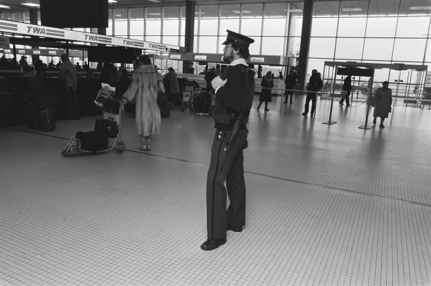 Polizeibeamter in Uniform mit Waffe in einem Flughafen-Terminal umgeben von Menschen und Gepäck, mit Gepäckwagen, Pfosten, Schildern, Deckenlampen und Glaswänden im Hintergrund.