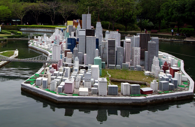 Ein Miniaturmodell einer Stadtlandschaft, umgeben von Wasser, mit Gebäuden, Grünflächen, einer Brücke, Menschen und einem Himmelhintergrund.