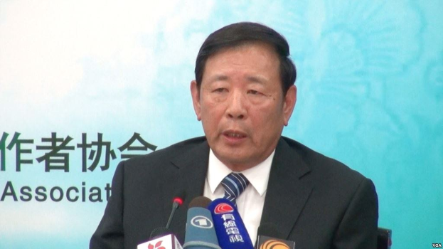 Ein Mann im Anzug sitzt an einem Tisch mit Mikrofonen und spricht zur Presse, mit einem Banner im Hintergrund.