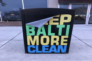 Schwarzer Müllcontainer mit dem Text "Keep Baltimore More Clean" auf einem Gehweg vor einem Gebäude mit Glastüren.