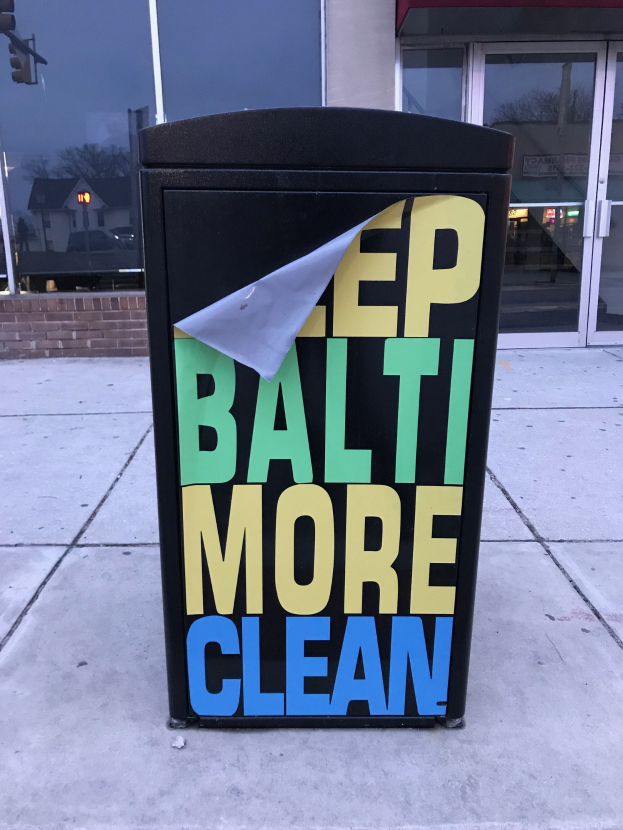 Schwarzer Müllcontainer mit dem Text "Keep Baltimore More Clean" auf einem Gehweg vor einem Gebäude mit Glastüren.