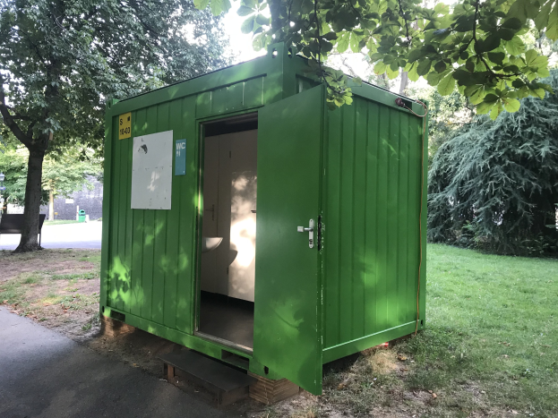Eine grüne tragbare Toilette mit einer Tür und einer angebrachten Tafel steht in einem Park, umgeben von Bäumen und einer Bank links, mit dem Himmel im Hintergrund.