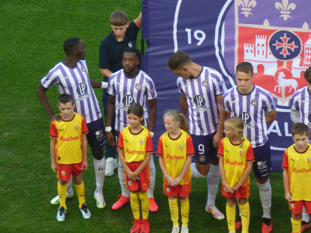 Gruppe junger Menschen auf einem saftig grünen Fußballfeld mit einem "Ligue 1"-Plakat im Hintergrund, einige tragen Trikots und andere kurze Hosen.