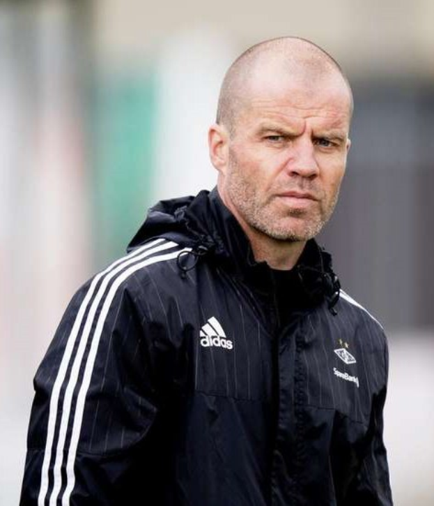 Ein kahlköpfiger Mann in einer schwarzen Jacke steht auf einem Fußballfeld und trägt eine marineblaue Jacke mit weißen Streifen und einem weißen Kragen, die Hände sind verschränkt und der Gesichtsausdruck ernst.