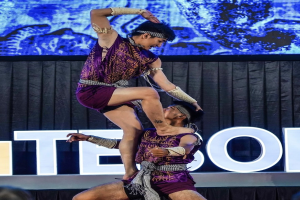 Zwei Akrobaten führen auf der Bühne eine Vorstellung auf, der Mann hält die Frau in seinen Armen, während beide purpurne Kleider tragen, mit Publikum und Bildschirm im Hintergrund.