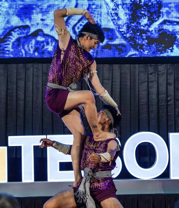 Zwei Akrobaten führen auf der Bühne eine Vorstellung auf, der Mann hält die Frau in seinen Armen, während beide purpurne Kleider tragen, mit Publikum und Bildschirm im Hintergrund.