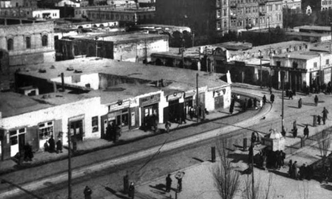 Schwarzes und weißes Foto einer Stadtstraße mit Fußgängern, umgeben von Gebäuden, Bäumen, Strommasten und Drähten.
