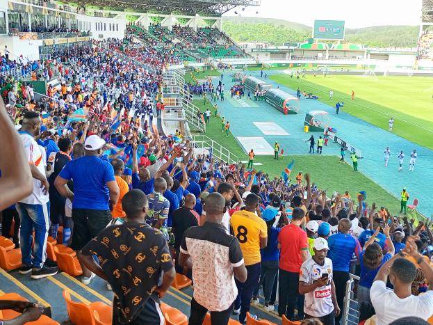Eine große Menschenmenge versammelt sich vor einem eingezäunten Fußballfeld unter einem klaren blauen Himmel mit Hügeln im Hintergrund und wartet gespannt auf ein Spiel während der Afrika-Cup 2019.