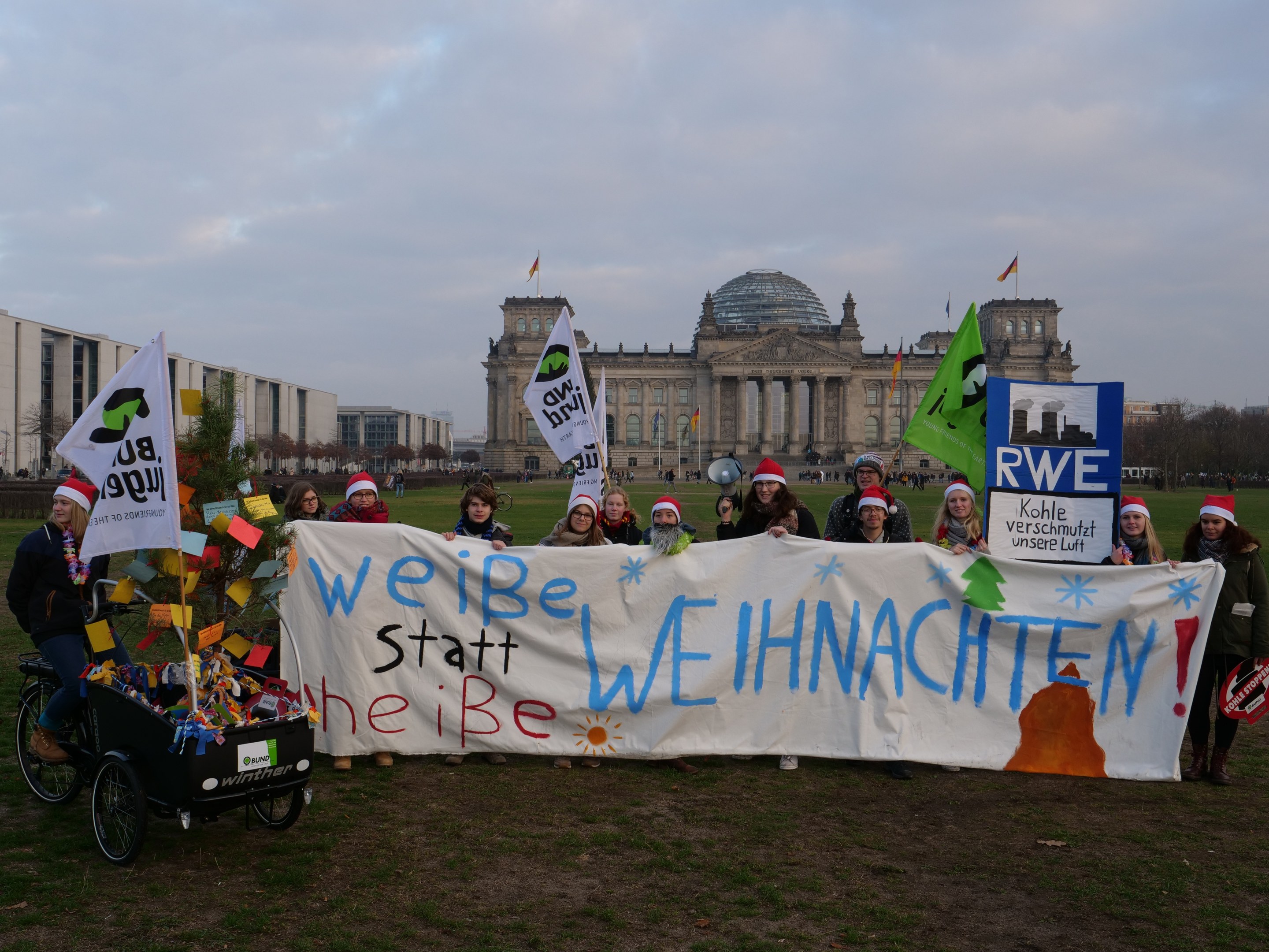Gruppe von Menschen in Mützen, die ein Banner vor dem Reichstaggebäude halten, mit einer Person auf einem Kinderwagen, Rasen, Bäumen, Gebäuden, Fahnen und bewölktem Himmel.