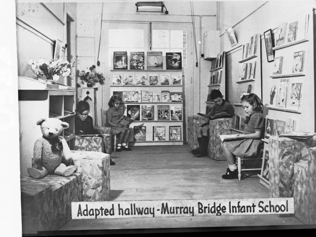 Eine Gruppe von Kindern sitzt auf Stühlen in einem Raum mit Regalen, einem Teddybär, einer Blumenvase und gerahmten Bildern an der Wand, mit dem Text "Adapted Hallway Murray Bridge Infant School" unten.