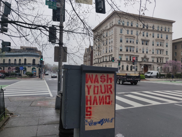 Eine belebte Stadtstraße mit Fahrzeugen, einem Fußgänger, einem "Waschen Sie Ihre Hände"-Müllcontainer, Verkehrszeichen, Schildern, einem Metallzaun, Grünflächen, Gebäuden und einem klaren Himmel.