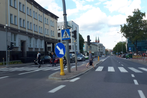 Stadtstraße mit Fußgängern an einer Ampelkreuzung, Fahrzeuge auf der Straße, Verkehrszeichen, Schilder, Laternenpfähle, Bäume, Gebäude mit Fenstern und ein bewölkter Himmel.