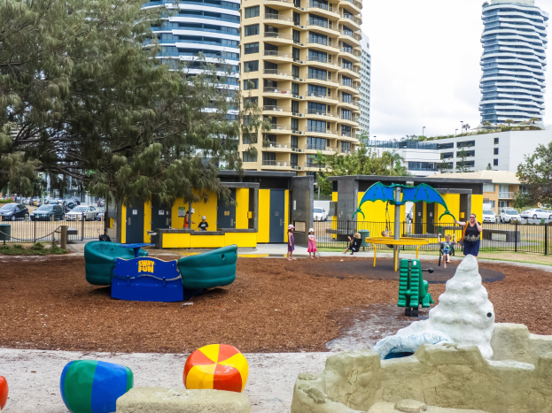 Spielplatz mit Kindern auf Geräten, umgeben von Bäumen und hohen Gebäuden, mit Fahrzeugen auf der Straße und sichtbarem Himmel darüber.