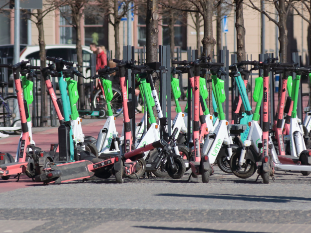 Eine Reihe von Elektro-Rollern, die an einer Straße geparkt sind, mit einer Person im Vordergrund, im Hintergrund sind Bäume und Gebäude zu sehen.