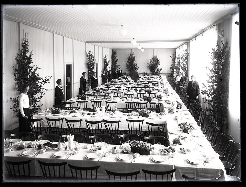 Festlich gedeckter Tisch mit langen Tischen, Stühlen, Geschirr und stehenden Gästen in einem Raum mit Pflanzen und Deckenbeleuchtung.