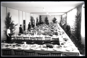 Festlich gedeckter Tisch mit langen Tischen, Stühlen, Geschirr und stehenden Gästen in einem Raum mit Pflanzen und Deckenbeleuchtung.