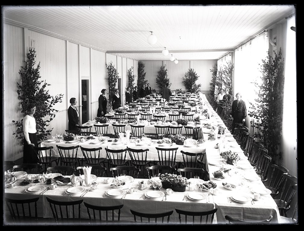 Festlich gedeckter Tisch mit langen Tischen, Stühlen, Geschirr und stehenden Gästen in einem Raum mit Pflanzen und Deckenbeleuchtung.