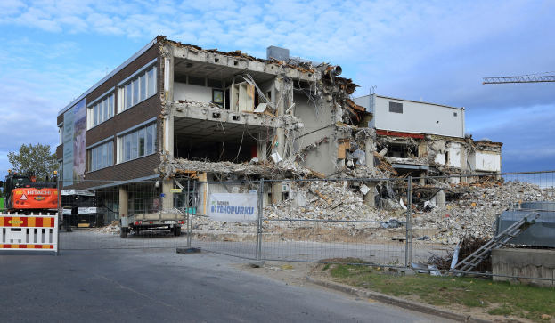 Ein großes Gebäude wird inmitten einer Straße abgerissen, umgeben von einem Metallzaun, Fahrzeugen, Gras, einem Schild, einer Fahne, Pfählen, einem Kran, Bäumen und einem bewölkten Himmel.