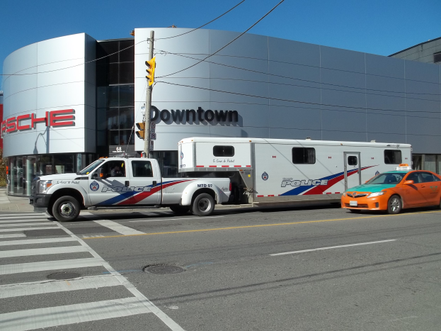 Ein Polizeiwagen steht vor einem Gebäude mit Text darauf, mit einer Verkehrsampel mit einem Mast und Drähten im Vordergrund und Bäumen und einem klaren blauen Himmel im Hintergrund.