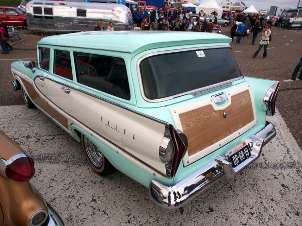 Ein gr├╝ner und wei├čer Kombi auf einem belebten Parkplatz mit Zelten, Masten, B├Ąumen und einem klaren blauen Himmel im Hintergrund.