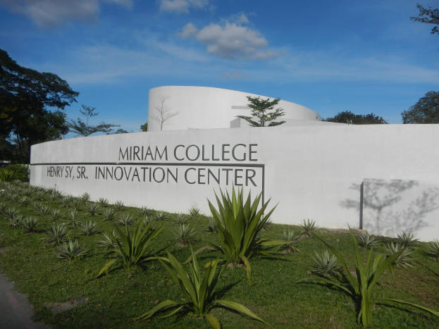 Außenansicht des Miriam College Innovationszentrums mit einer textbedeckten Wand, umgeben von Grün, einem Weg, Bäumen und einem bewölkten Himmel.