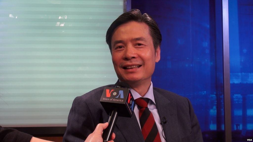 Ein Mann in Anzug und Krawatte hält ein Mikrofon, mit der Hand eines Reporters sichtbar, der vor einem Fenster und einer Wand spricht.