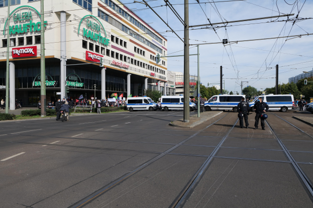Polizeibeamte stehen auf einer Straße neben einer Tram mit Fahrzeugen, Strommasten, Gebäuden, Bäumen und einem bewölkten Himmel im Hintergrund.