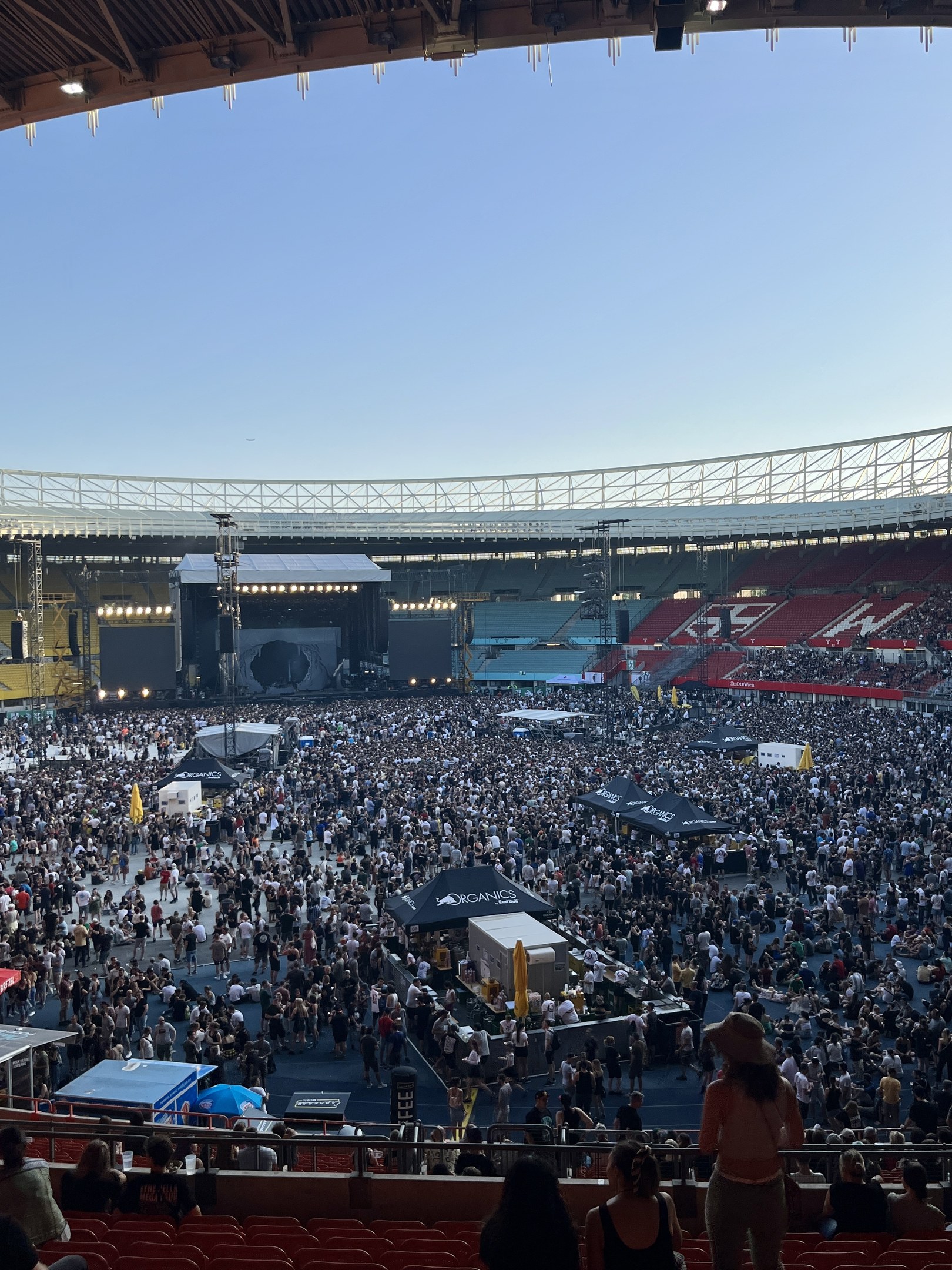 Große Menschenmenge auf Sitzplätzen und Stehplätzen in einem Stadion mit Zelten, Geländern und Überdachung, vor einem Himmel als Hintergrund im O2 Arena in London.
