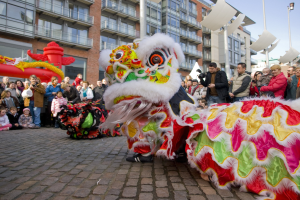 Ein lebendiges chinesisches Neujahrsfest in Amsterdam mit einer Löwen-Tanz-Show im Vordergrund und einer Menschenmenge, einige halten Kameras, vor einem Hintergrund aus Gebäuden, Laternenmasten und einem klaren blauen Himmel.