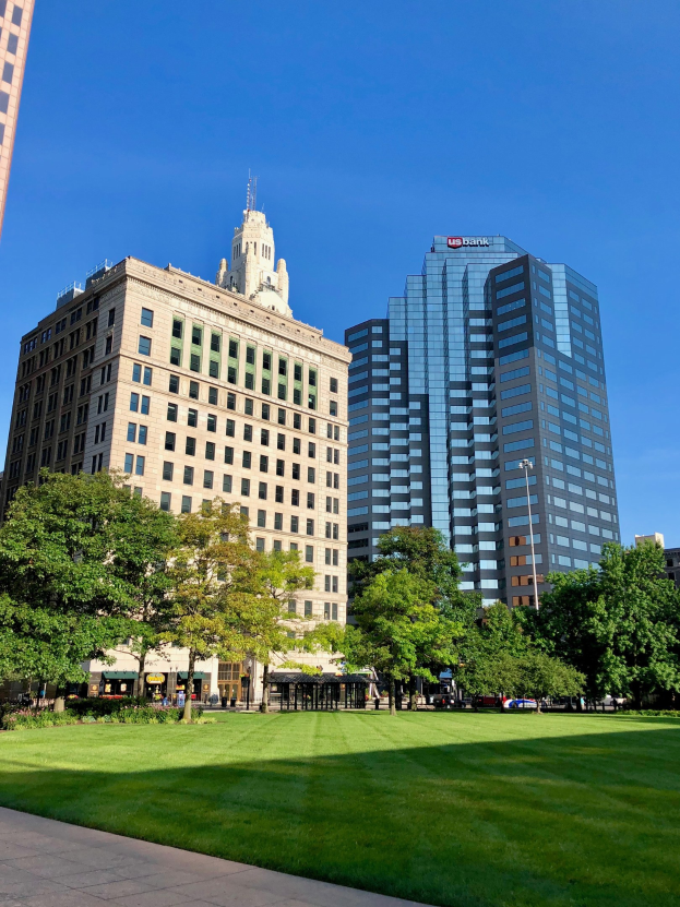 Stadtpark mit hohen Gebäuden im Hintergrund, umgeben von grünem Gras, Bäumen und Laternenmasten, mit Menschen, die spazieren gehen und dem Himmel darüber.