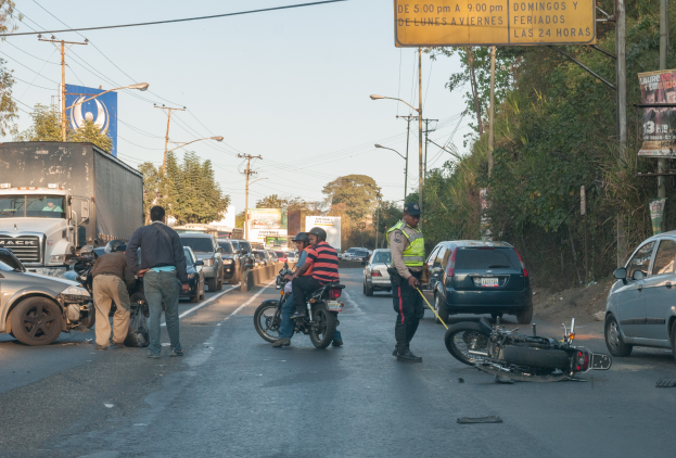 Eine Gruppe von Menschen um ein verunglücktes Motorrad auf der Straße herumstehend mit mehreren Fahrzeugen, darunter ein Lastwagen, und einem Hintergrund aus Bäumen, Polen, Lampen, Schildern und Himmel.