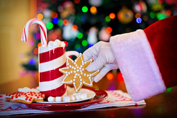 Eine Person mit Santa Claus Hut hält einen Becher heiße Schokolade und einen Bonbonstab und hat Plätzchen und ein Tuch auf dem Tisch, vor einem Weihnachtsbaum und einer Wand.