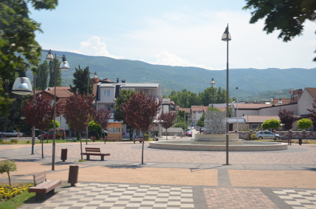 Ein Stadtplatz mit einem zentralen Brunnen, umgeben von Bänken, Laternen, Bäumen, Pflanzen, Blumen und Gras, mit Gebäuden, Fahrzeugen, Hügeln und einem bewölkten Himmel im Hintergrund.