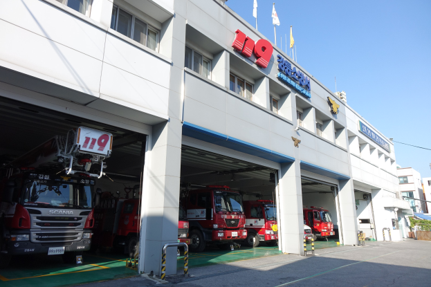 Feuerwache mit mehreren Feuerwehren vor dem Gebäude geparkt, mit Fenstern, Säulen, Namensschildern und Flaggen im Vordergrund auf Stangen, vor einem Hintergrund aus Gebäuden und einem klaren blauen Himmel.