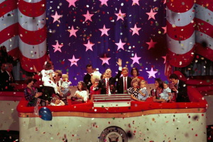 Eine Gruppe von Menschen auf einer Bühne mit einem Rednerpult, das ein Logo, Luftballons und Konfetti im Hintergrund zeigt, mit Sternen und einem Vorhang im Hintergrund.