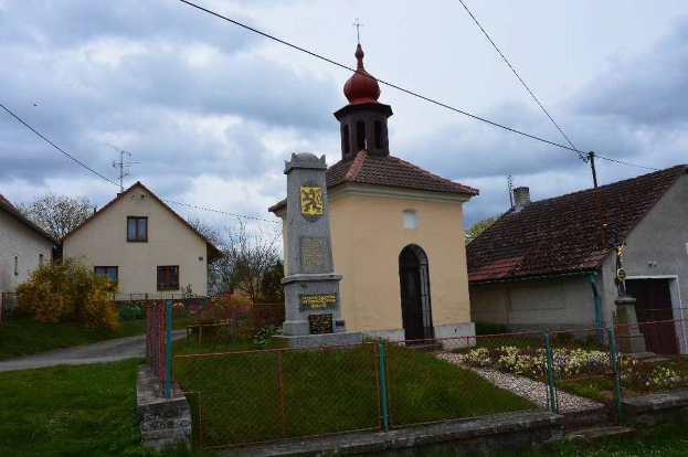 Kleine steinerne Kirche namens Kirche des Heiligen Grabes mit einem Kirchturm, umgeben von Wohnhäusern, Grünflächen, einem Zaun und einem bewölkten Himmel.