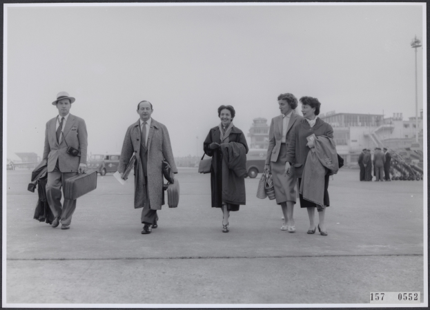 Eine Gruppe von Menschen öffnet den Weg über das Rollfeld eines Flughafens, jeder von ihnen trägt einen Koffer, mit Fahrzeugen, Gebäuden, Masten und einem klaren blauen Himmel im Hintergrund.