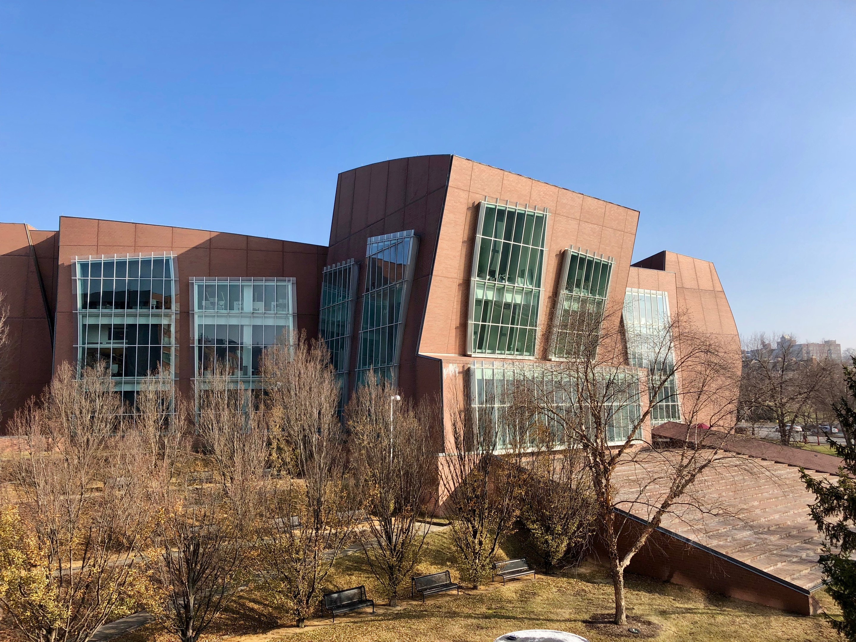 Großes mehrstöckiges Gebäude mit zahlreichen Fenstern, umgeben von Bäumen, Bänken, Gras und einem Laternenmast, mit anderen Gebäuden im Hintergrund unter einem klaren blauen Himmel.