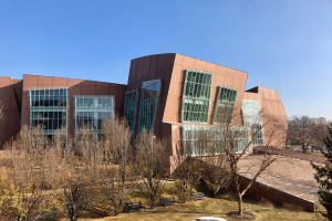 Großes mehrstöckiges Gebäude mit zahlreichen Fenstern, umgeben von Bäumen, Bänken, Gras und einem Laternenmast, mit anderen Gebäuden im Hintergrund unter einem klaren blauen Himmel.