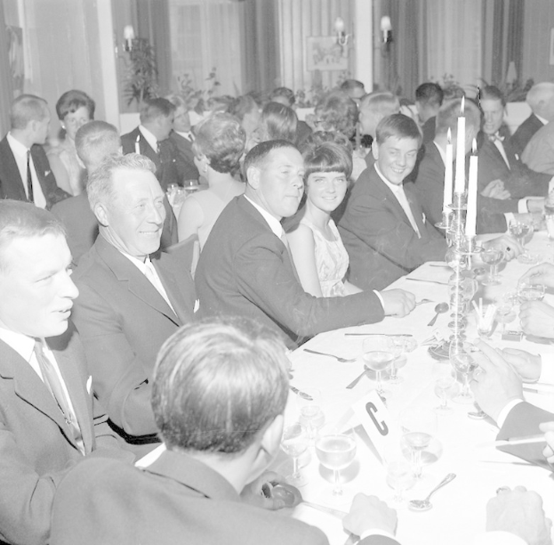 Eine Gruppe von Menschen sitzt um einen langen Tisch, lacht und genießt die Gesellschaft des anderen. Auf dem Tisch befinden sich Gläser, Bestecke, Kerzenhalter und andere Gegenstände. Im Hintergrund sind Vorhänge und Wandlampen zu sehen, die eine gemütliche Atmosphäre schaffen.