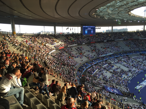 Eine große Menschenmenge sitzt im Allianz Arena Stadion in München, Deutschland, und schaut ein Fußballspiel, mit einer Bühne auf der rechten Seite, Fahnen, Stangen und einem Bildschirm im Hintergrund und dem Himmel oben sichtbar.