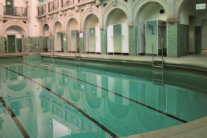 Indoor-Schwimmbad mit klarem Wasser, umgeben von gefliestem Boden, großes Gebäude mit Bögen, Säulen, Geländern, Türen und Tafeln mit Text.