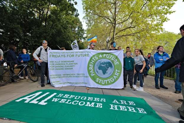 Gruppe von Menschen mit einem Banner, auf dem "Alle Flüchtlinge sind hier willkommen" und einer Fahne, mit Fahrrädern, einem Zaun, einer Straßenlaterne, einem Schild, Bäumen und einem bewölkten Himmel im Hintergrund.